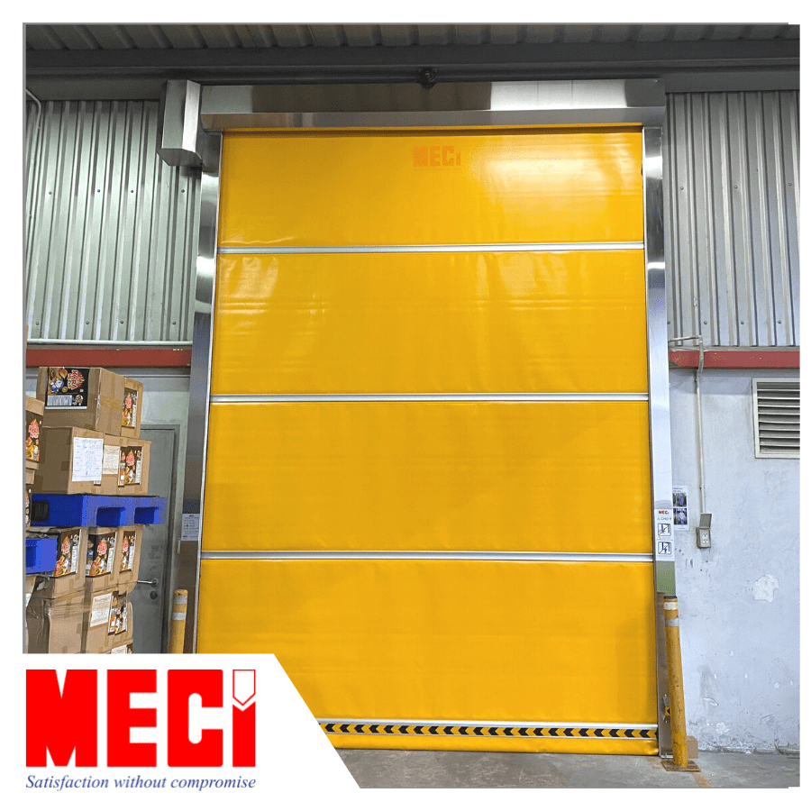 Cửa cuốn nhanh PVC màu vàng không có cửa sổ, lắp tại cửa ra vào kho. Bên trái có thùng hàng chứa sản phẩm. 