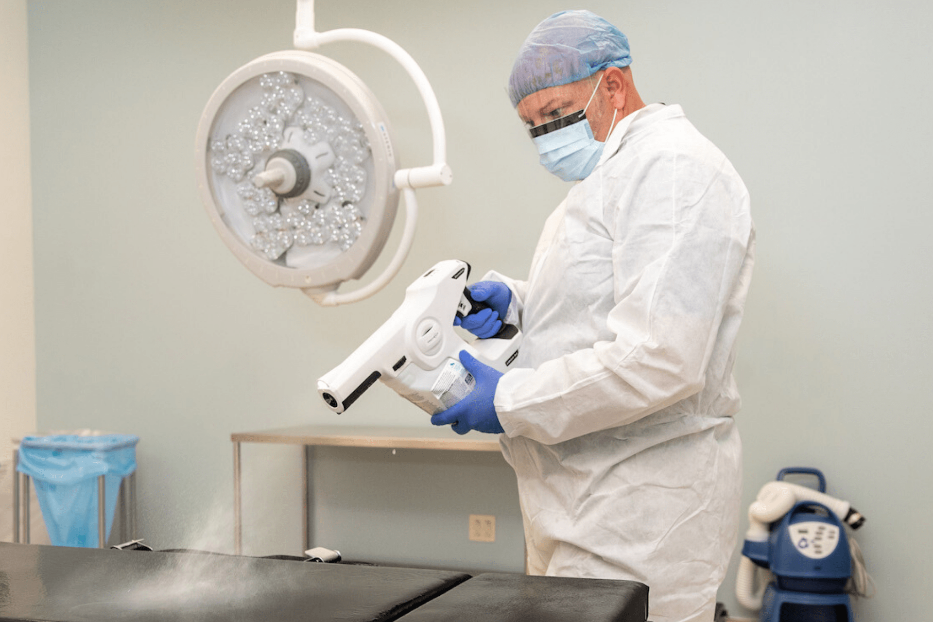 Người đàn ông mặc đồ bảo hộ trắng, găng tay vàng đang dùng vòi xịt xịt vào bề mặt đồ vật.