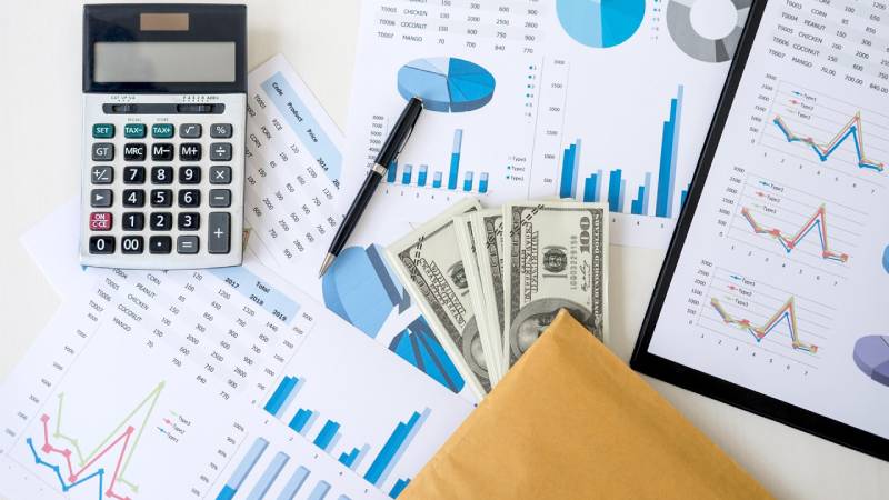 Trên bàn có báo cáo doanh thu, báo cáo chi phí quản lý doanh nghiệp, máy tính và tiền