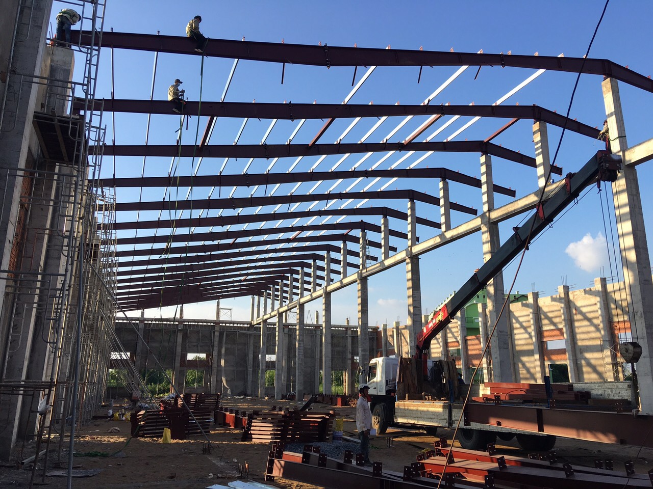 Nhà xưởng đang được thi công có 2 người đàn ông đang ở trên sườn mái và 1 người phía dưới.