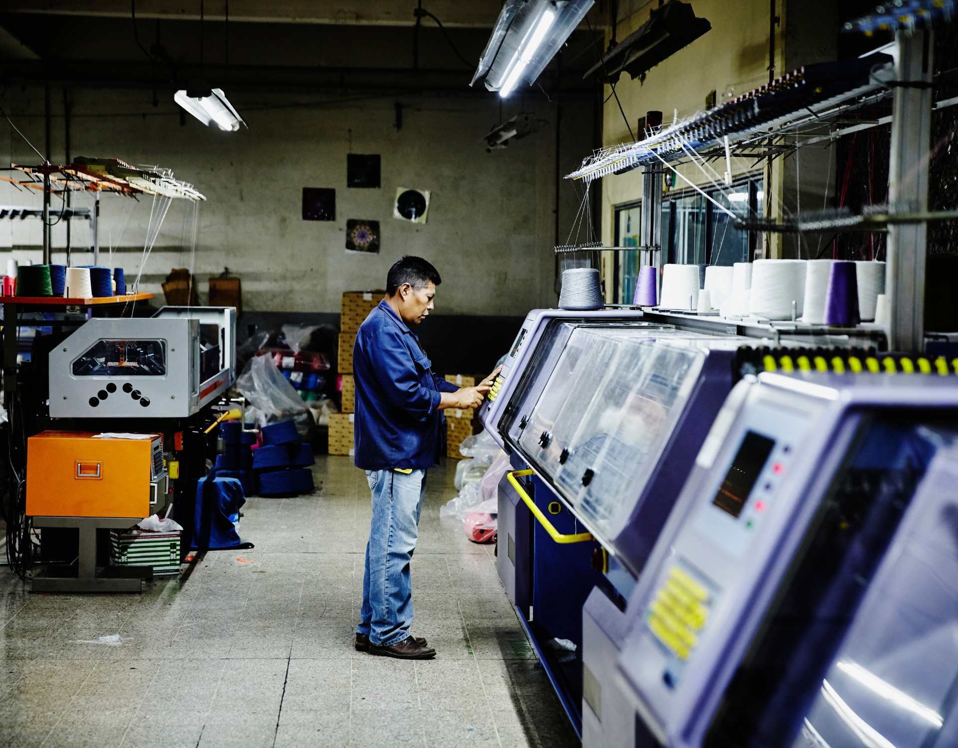 một người nam đnag điều khiển máy dệt vải, xung quanh là nhà xưởng với nhiều máy móc khác