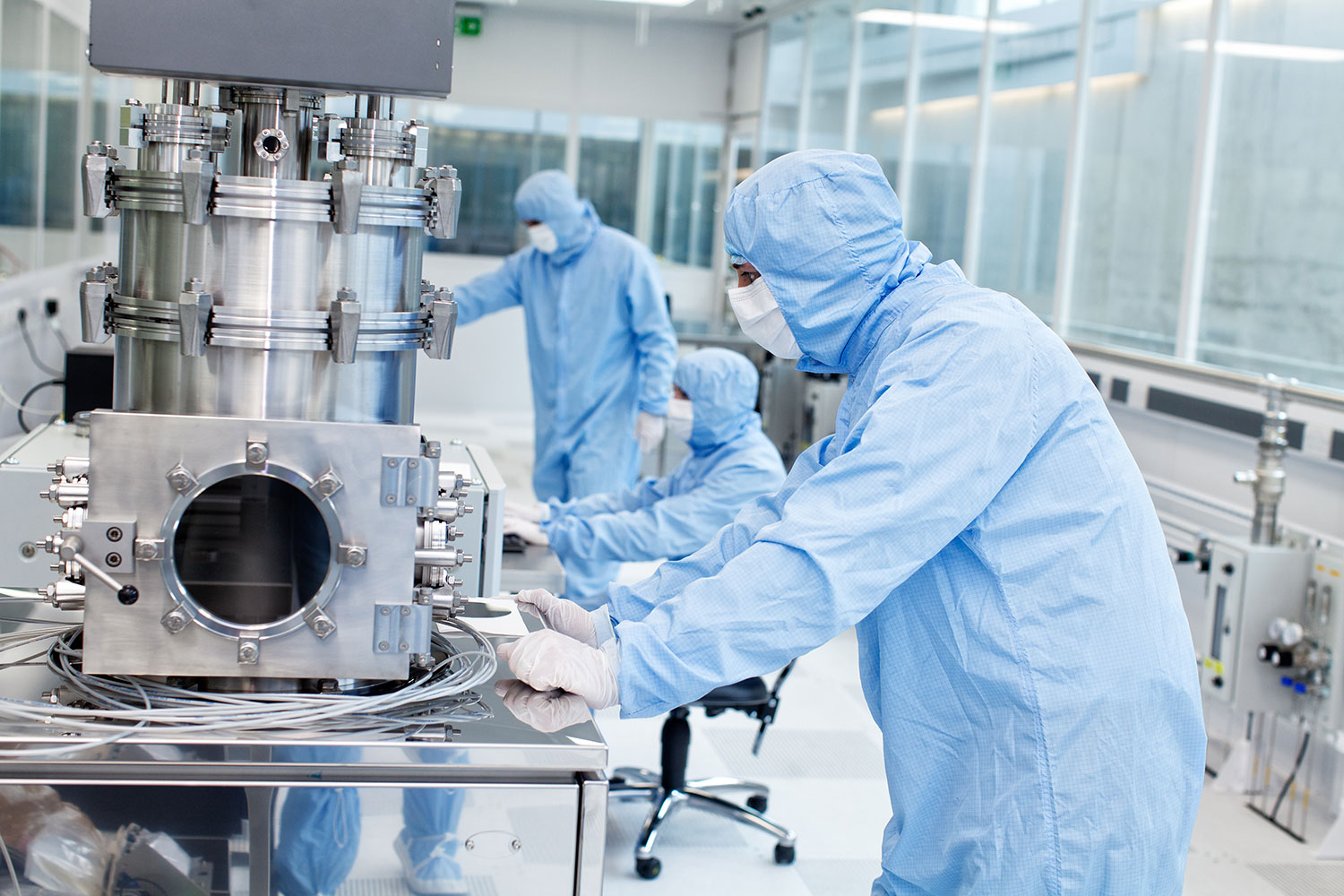 3 người mặc quần áo phòng sạch màu xanh nhạt đang làm việc với nhiều thiết bị máy móc