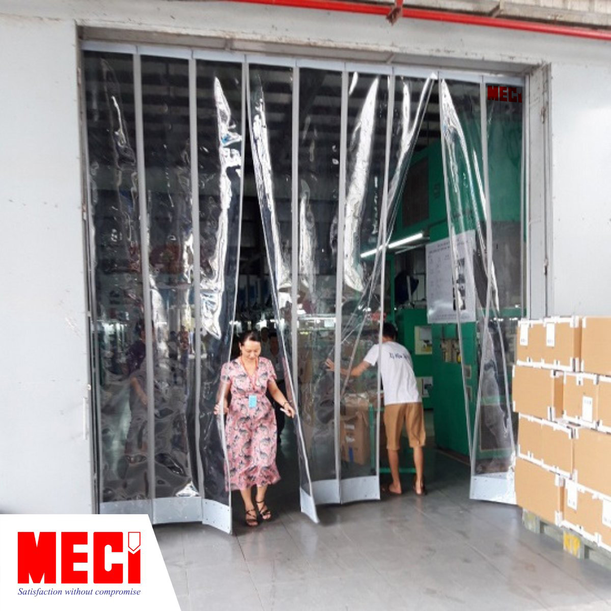 một nguời phụ nữ từ trong đi ra, và 1 người đàn ông đẩy xe hàng đi vào ngang qua cái rèm nam châm được lắp ở lối ra vào nhà xưởng