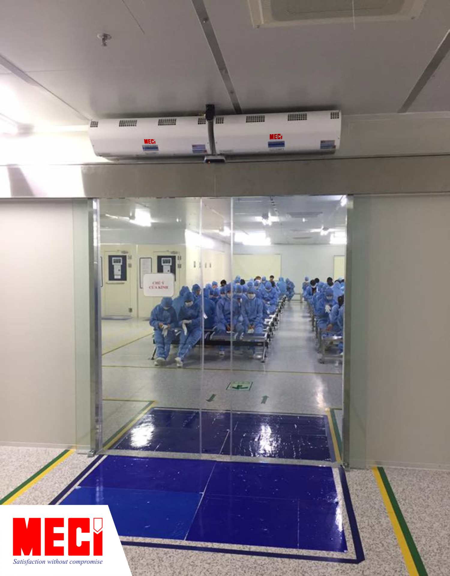 hai chiếc quạt cắt gió lắp phía trên lối ra vào có cửa kính dạng lùa, bên ngoài có nhiều người mặc đồ bảo hộ phòng sạch đnag ngồi chờ