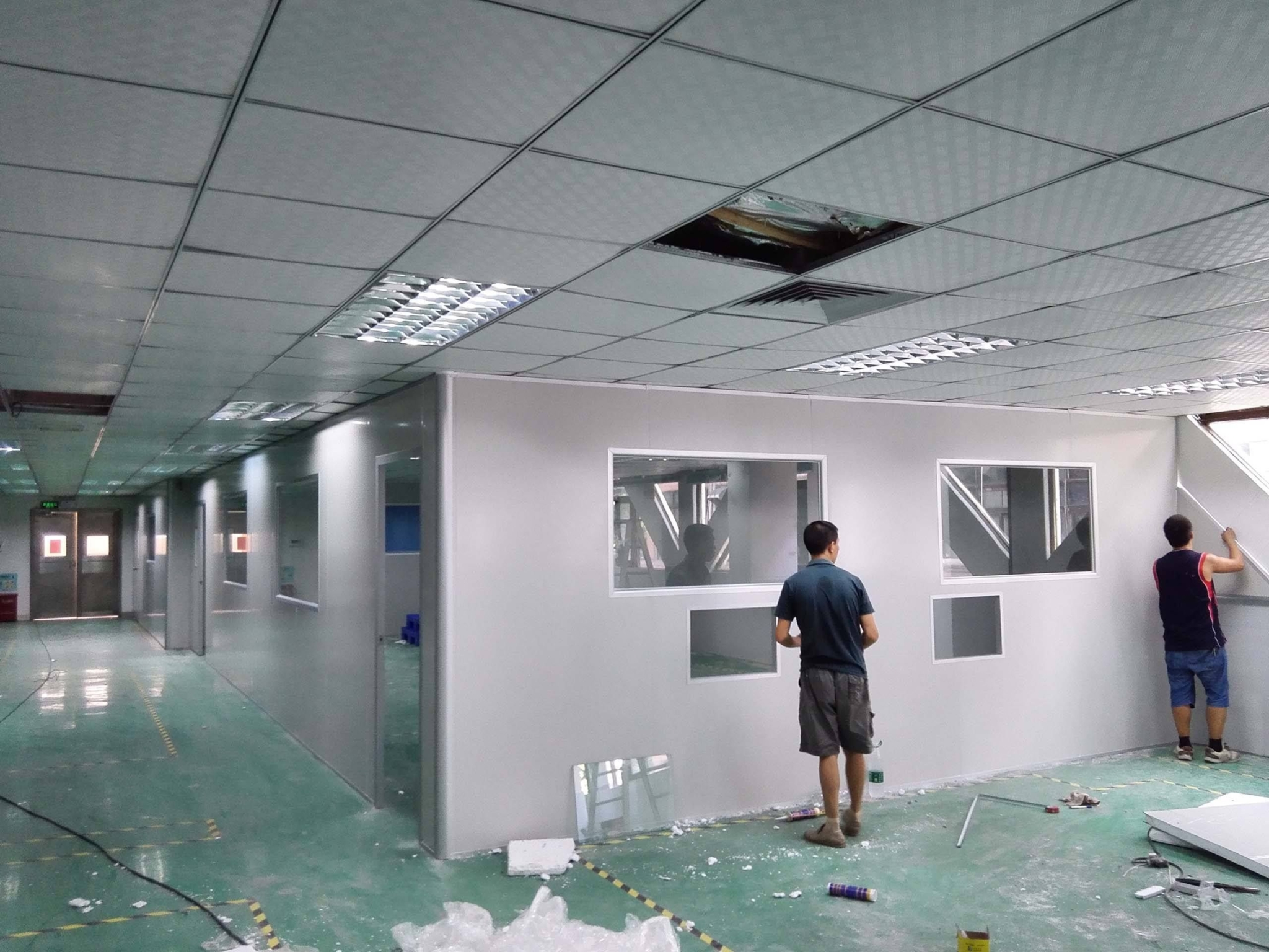 Có 2 người đàn ông đang thi công phòng sạch. Người áo xanh đang nhìn về phía người áo đen. Sàn nhà có màu xanh, vách tường màu trắng, trần nhà có những ô trống chưa được lắp đặt.