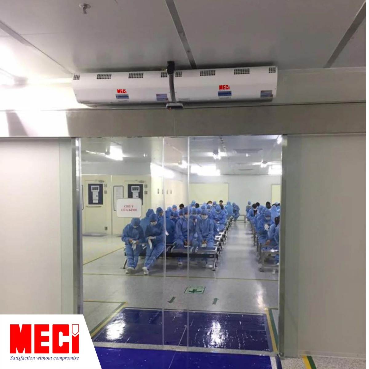 hai chiếc quạt cắt gió lắp phía trên lối ra vào có cửa kính dạng lùa, bên ngoài có nhiều người mặc đồ bảo hộ phòng sạch đang ngồi chờ