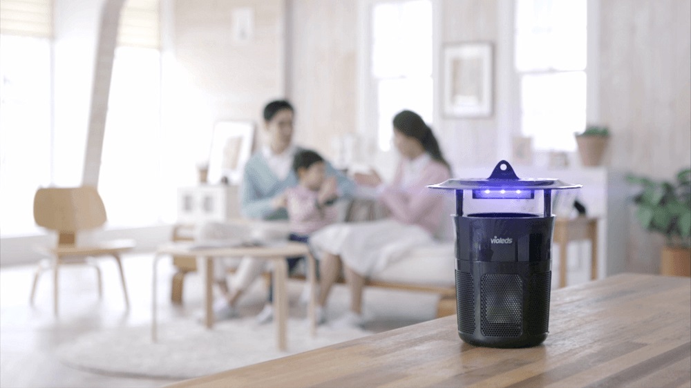 đèn bắt muỗi được đặt trên bàn, background phía sau là một gia đình đang sinh hoạt