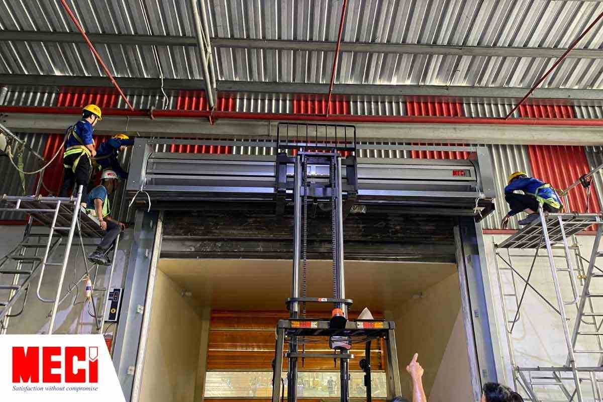 Xe nâng nâng cửa cuốn nhanh nan nhôm lên cao, hai bên là các dàn giáo có kỹ thuật MECI ngồi lên trên
