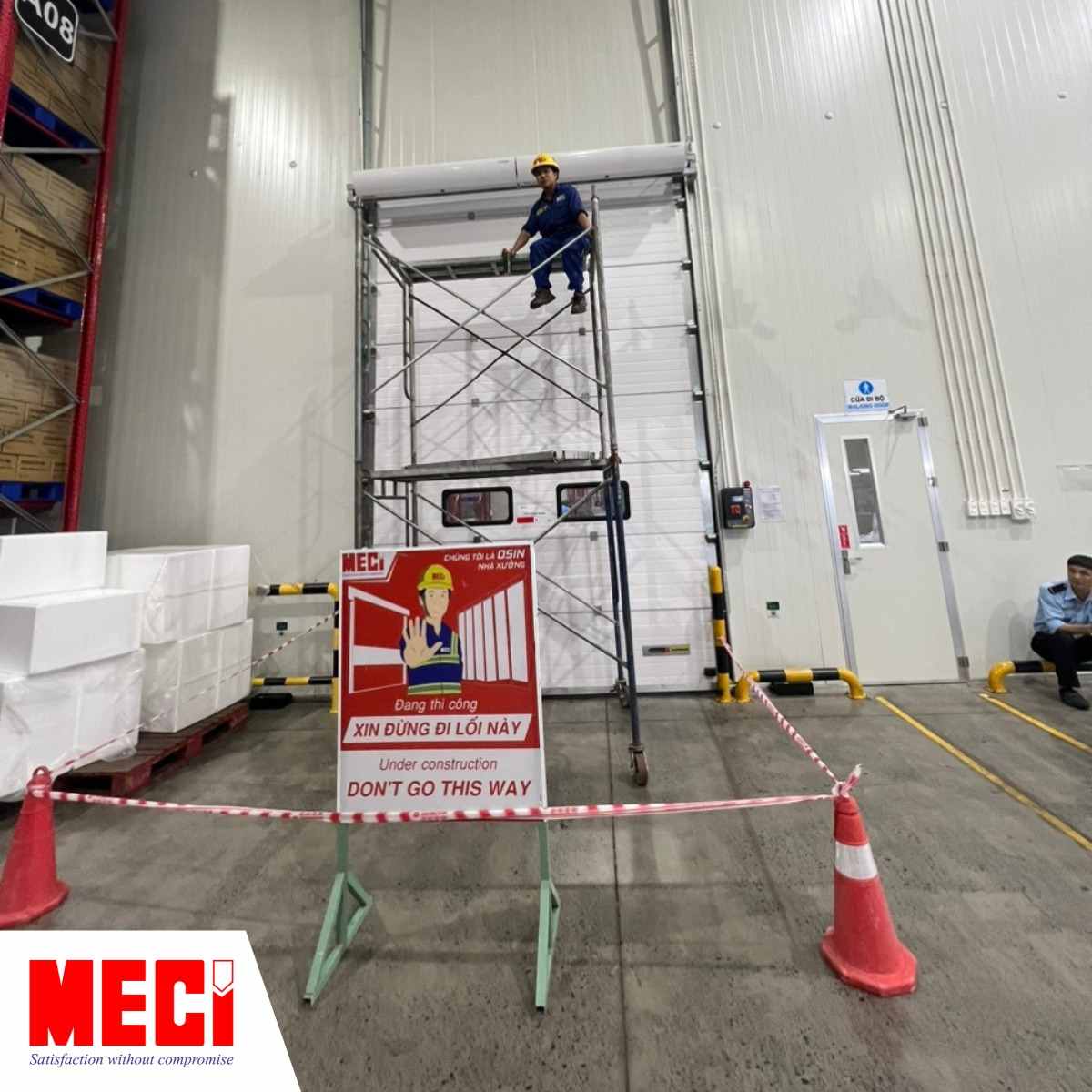 Nhân viên MECI đang ngồi trên dàn giáo, phía sau là cửa trượt trần, phía trước là bảng hiệu thi công