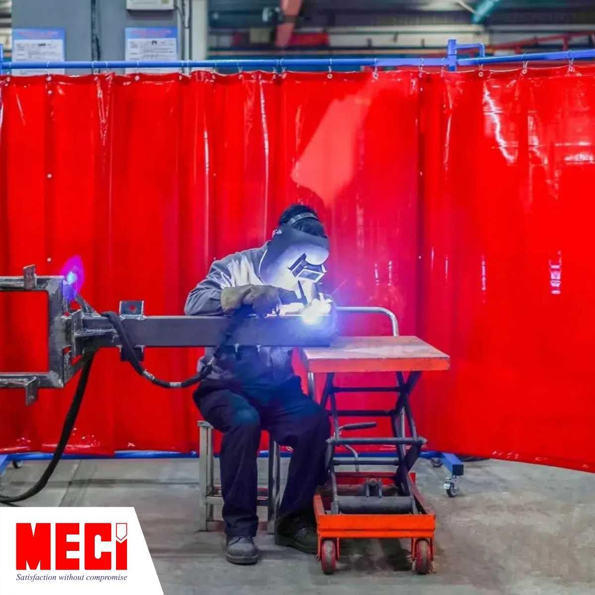 một người công nhân đang ngồi hàn sắt, phía sau là rèm nhựa kéo dạt ngăn tia lửa hàn