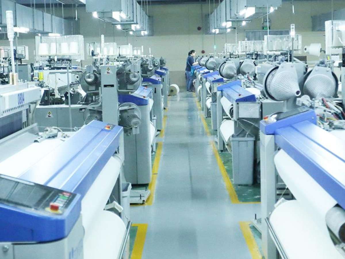 khung cảnh bên trong nhà máy dệt may, cò hàng máy móc đang hoạt động và các nhân viện đứng kế bên một vài máy